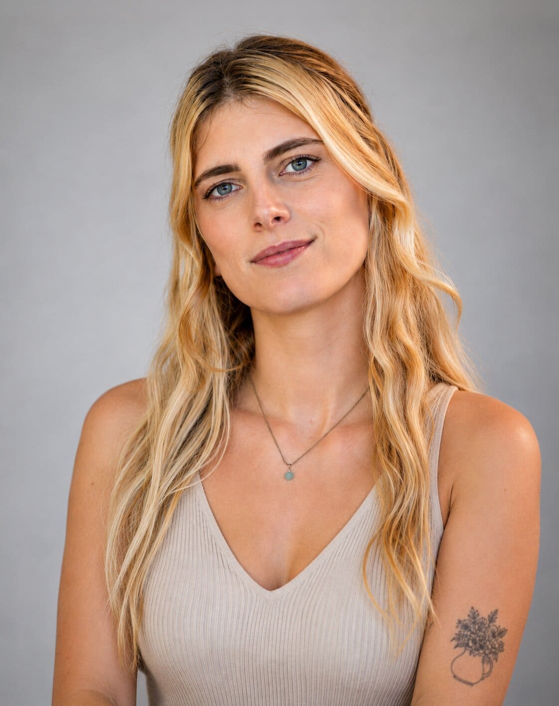 A smiling woman with long blonde hair, blue eyes, and a flower vase tattoo on her left arm.