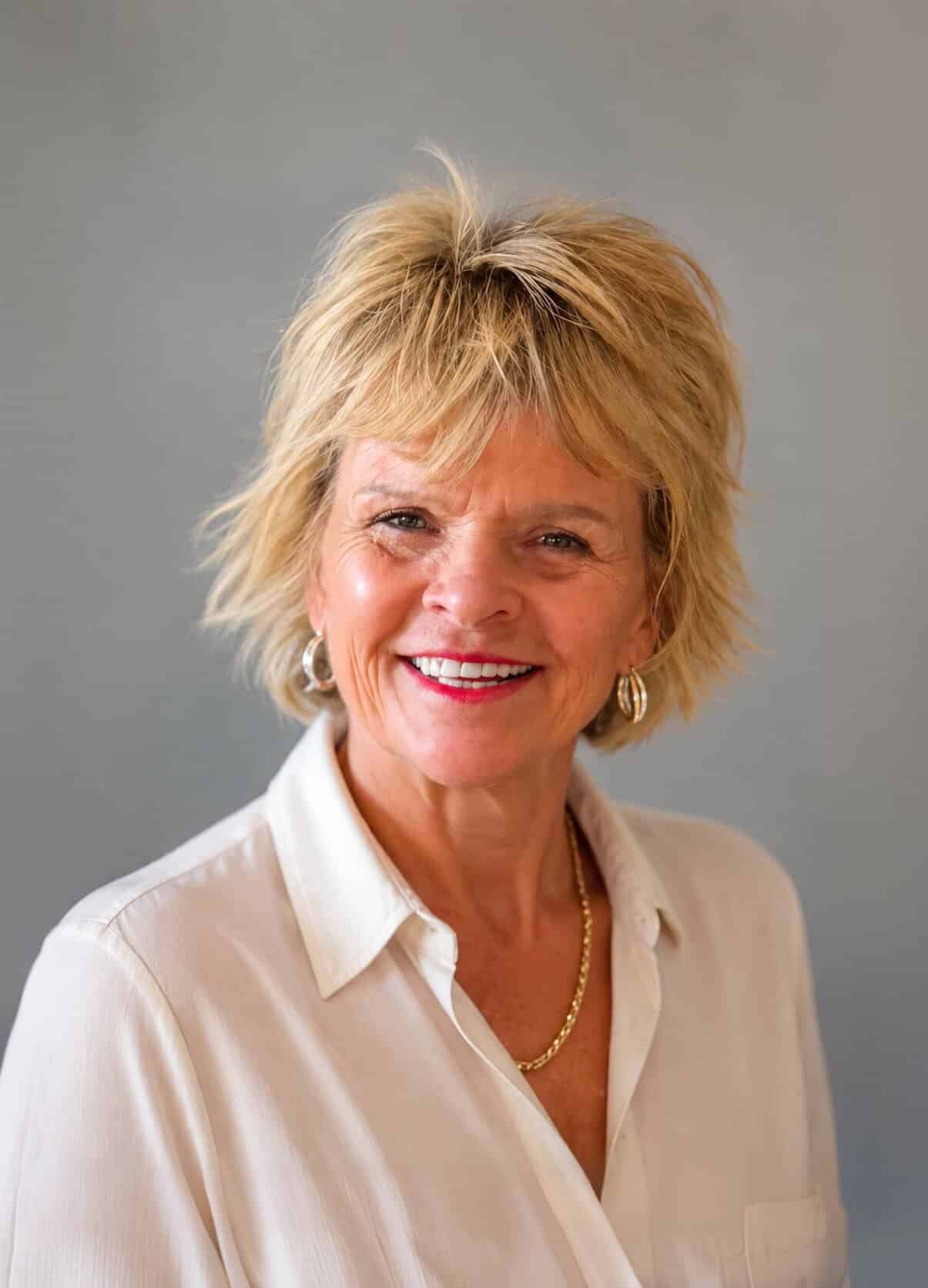 A smiling woman with short blonde hair, a cream shirt, and a gold necklace looks directly forward.