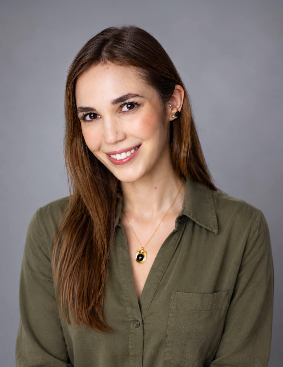 A smiling woman with long brown hair wears a green button-up shirt and a gold necklace with a black stone.