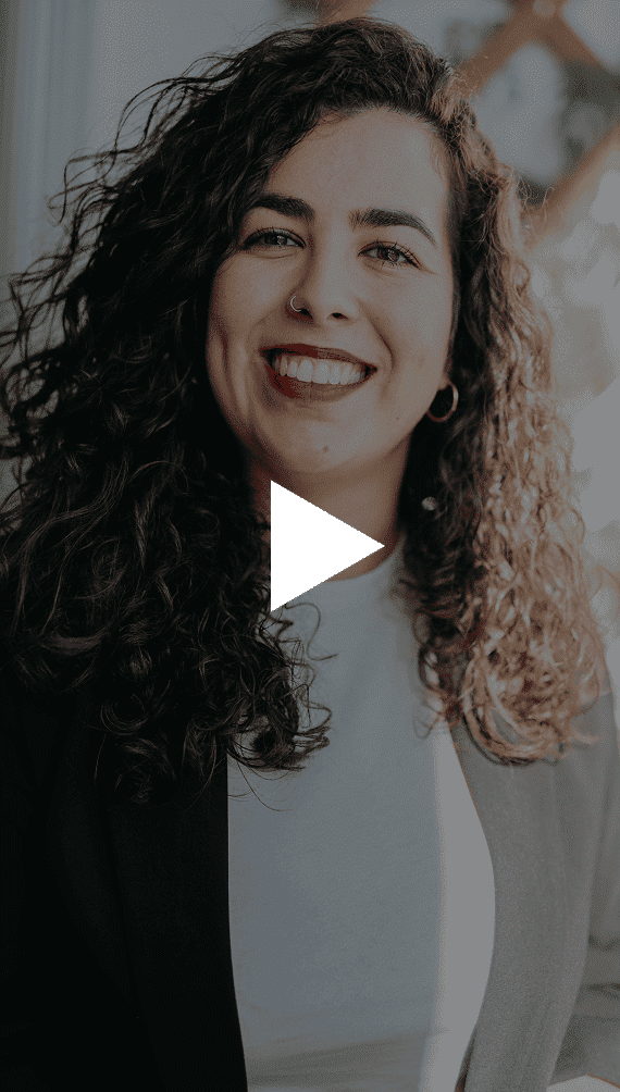 A smiling woman with dark curly hair, a nose ring, and a white play button overlay.