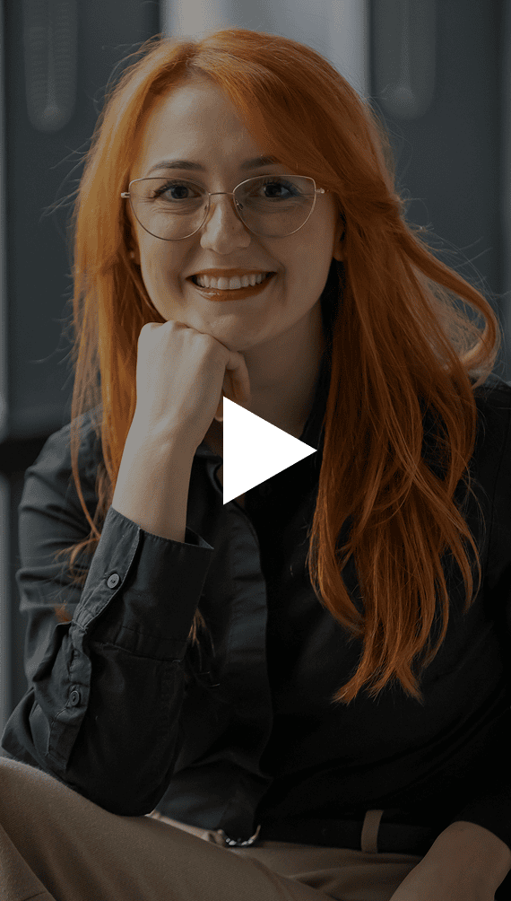 A smiling woman with long red hair and glasses rests her chin on her hand, with a white play button overlay.