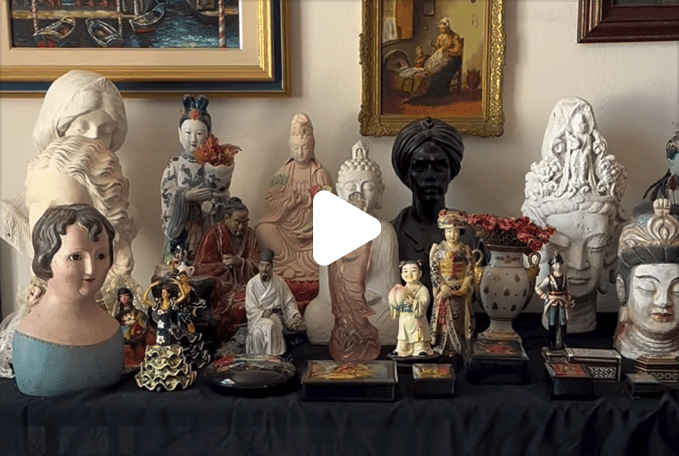 A collection of diverse statues and busts, including a black bust with a turban, on a dark cloth with paintings above.