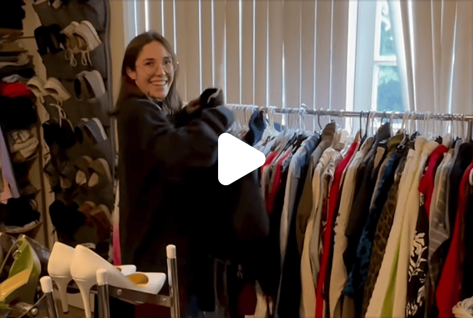 A smiling woman stands in a room with a shoe organizer, a clothing rack, and white high heels.