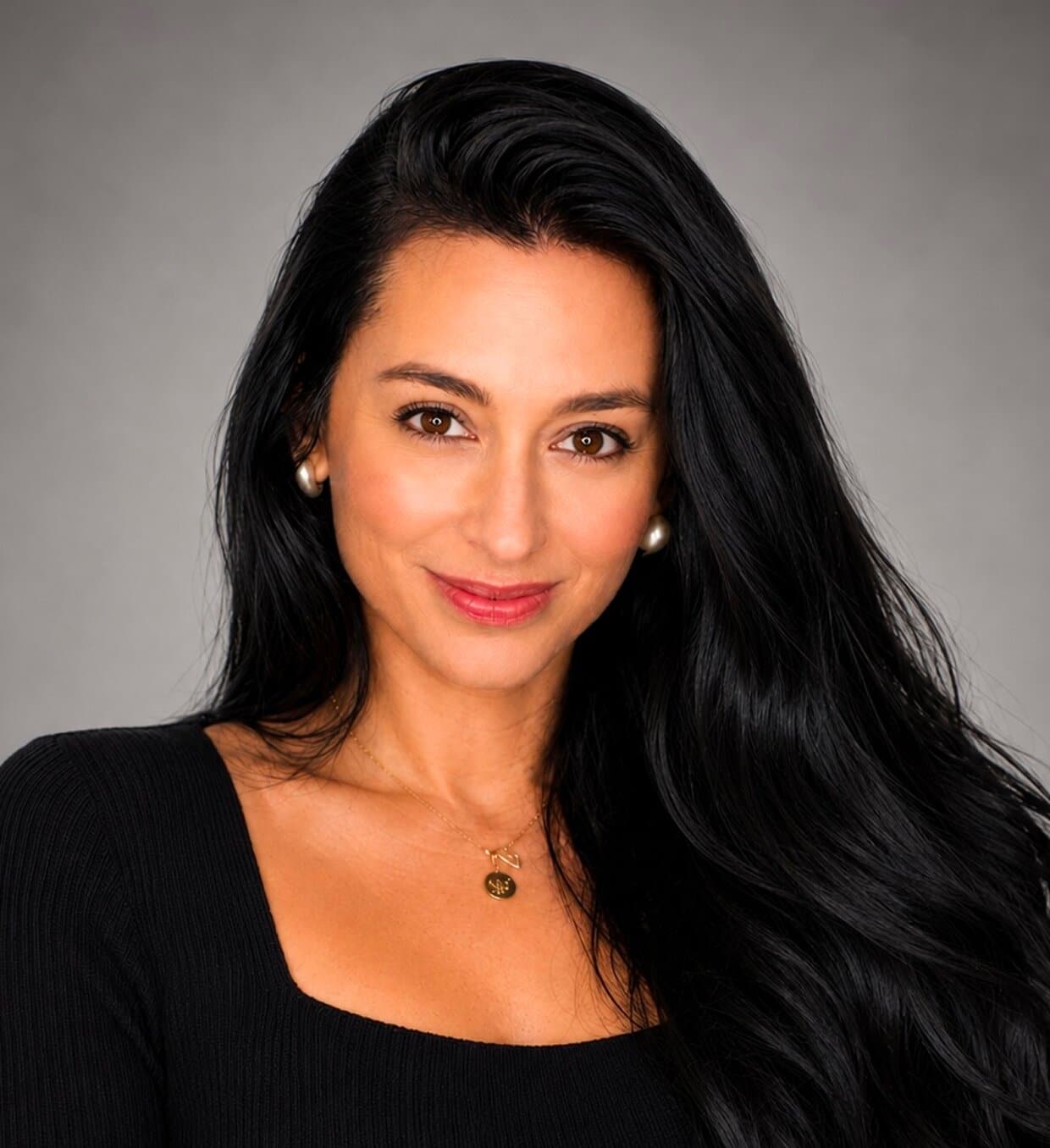 A smiling woman with long dark hair, light brown eyes, and fair skin wears a black top and gold necklace.