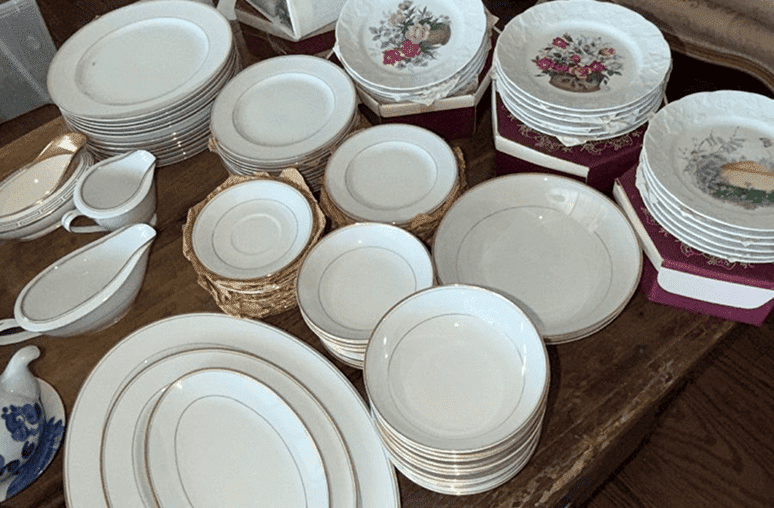 A wooden table is covered with stacks of white plates, some with gold rims and some with floral patterns.