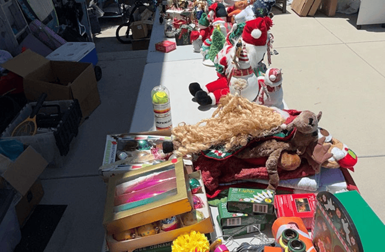 A long table is covered with various items for sale, including Christmas decorations, toys, and a "DUNLOP" tennis ball container.