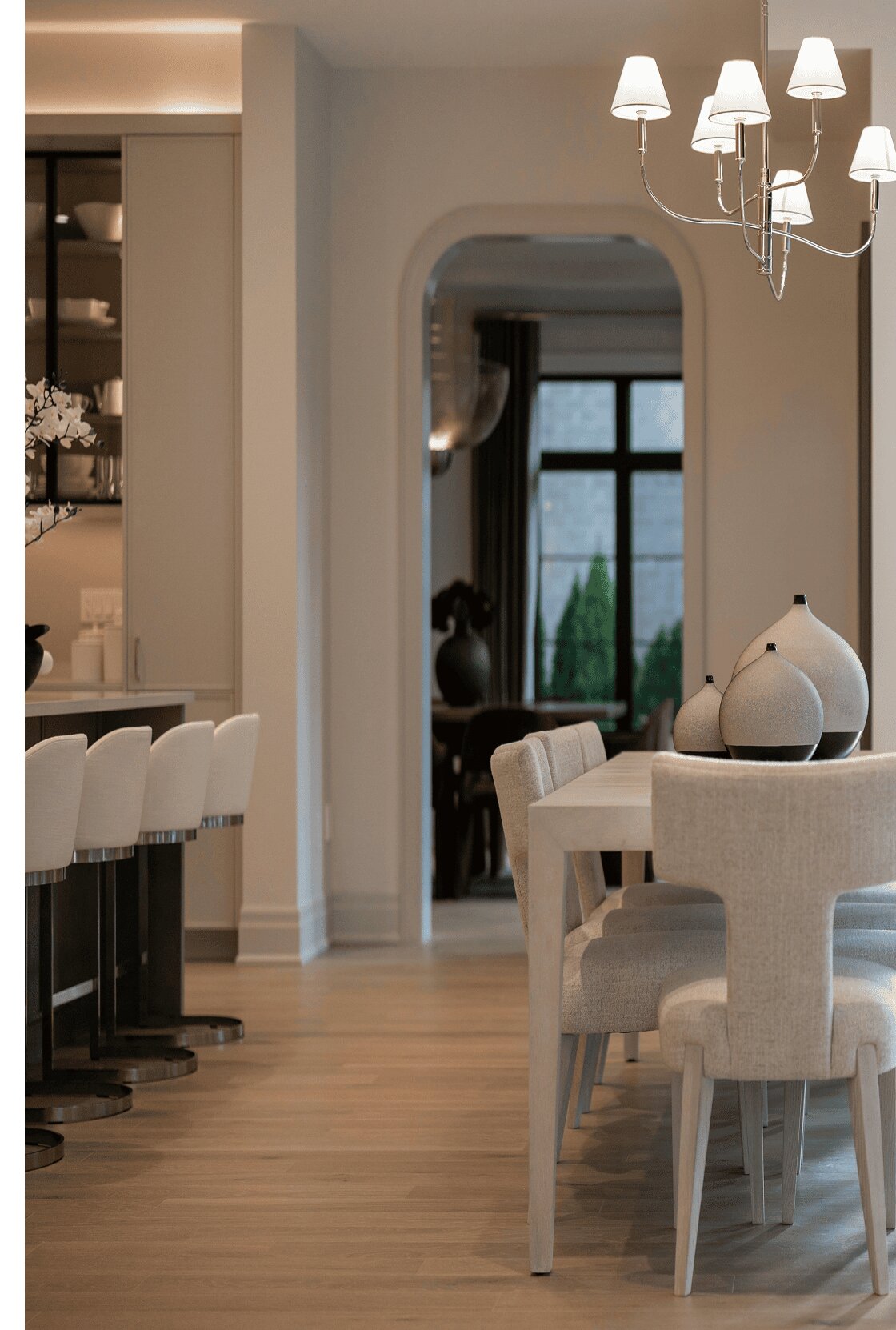 A modern dining room with light wood floors, a white table, chairs, and a chandelier, leading to a kitchen.