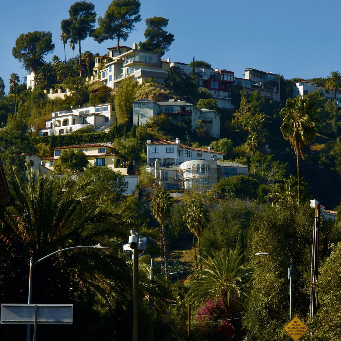 Numerous multi-story houses and lush trees cover a steep hillside under a clear blue sky.