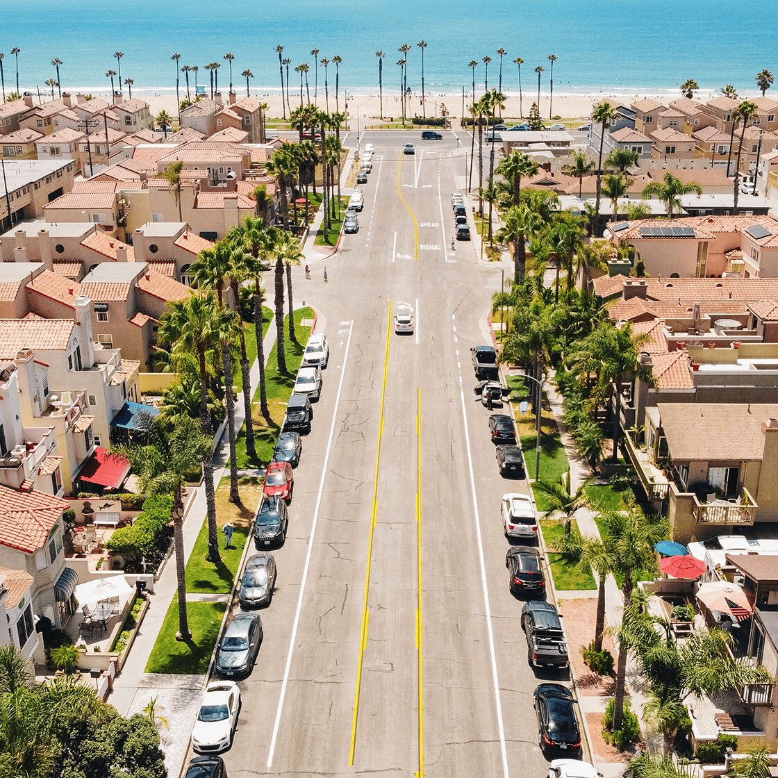 An aerial view of a beach town with palm trees, houses, and cars parked along a street leading to the ocean.